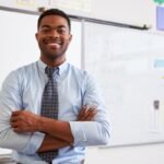 Professor sorridente com braços cruzados em sala de aula com quadro branco